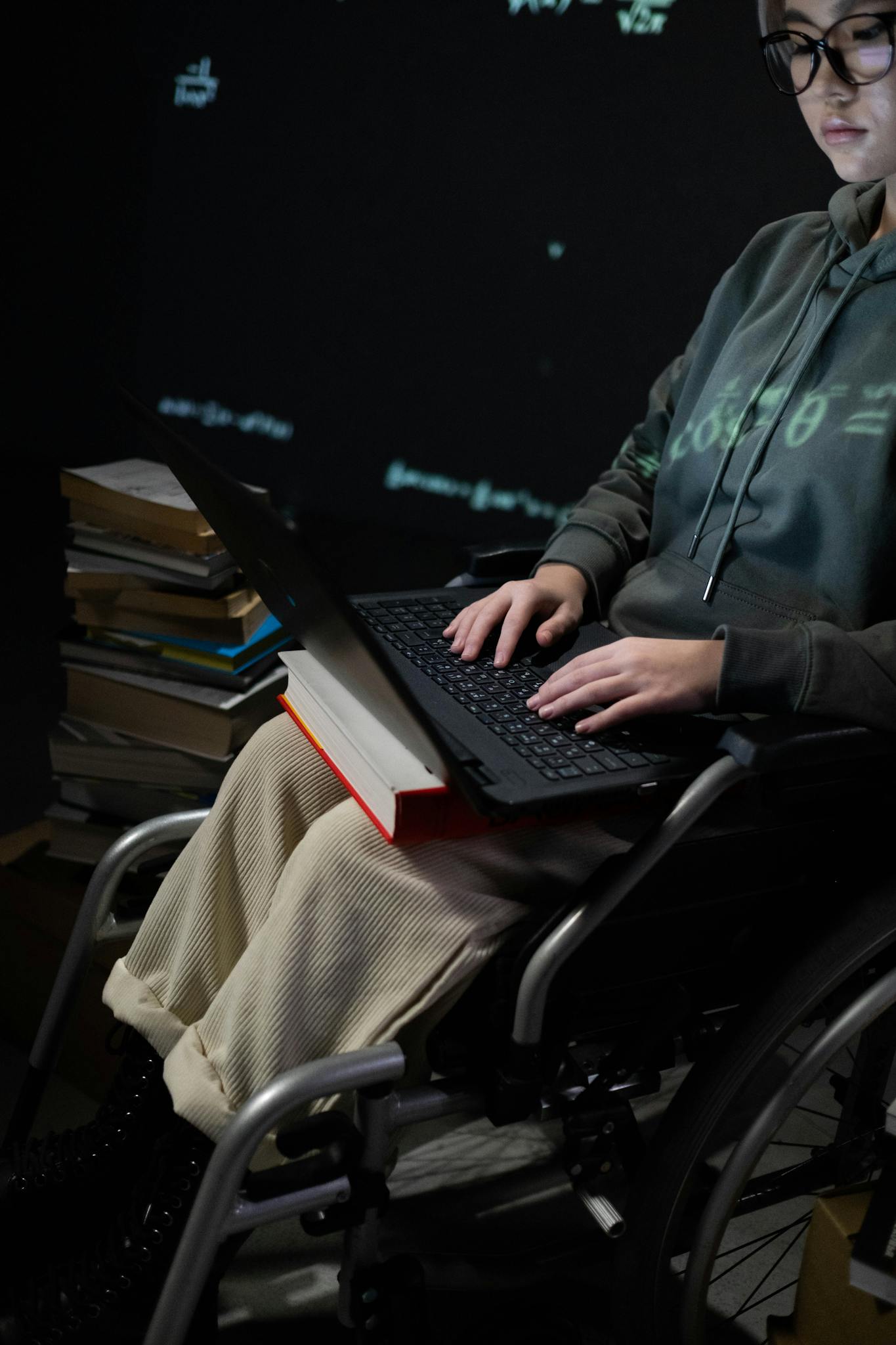 A teenager in a wheelchair typing on a laptop, surrounded by books, suggesting studying.